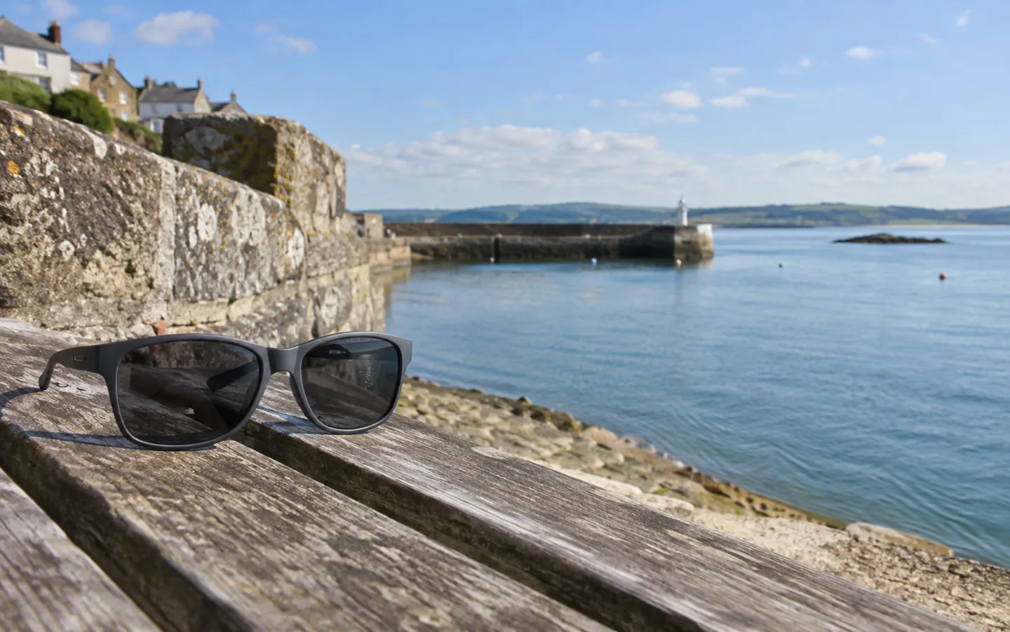 A pair of grey-lensed sunglasses resting on a weathered wooden bench by a sunny harbour wall, with calm sea behind